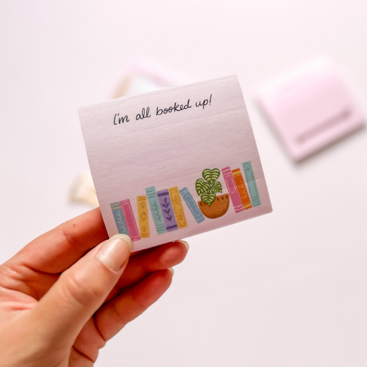 Hand-illustrated pink sticky notepad featuring a row of colorful books and a potted plant, with the label 'I'm All Booked Up'.